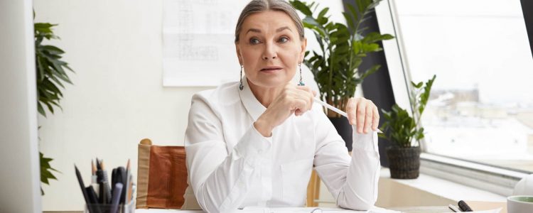 Attractive experienced 50 year woman chief architect with gray hair studying drawings on desk in front of her, making notes and comparing date with measurements on computer, having focused look