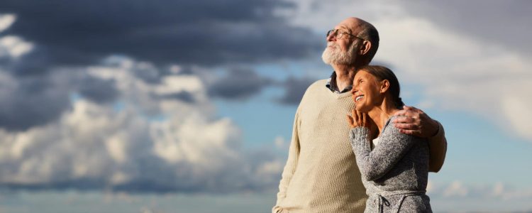 Amorous seniors standing by water against stormy sky and enjoying sundown