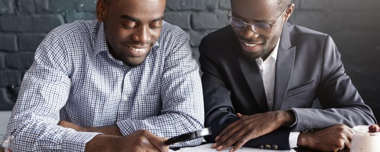 Happy African American office workers dressed in formal clothing having cheerful looks, studying and amalyzing legal documents on table using magnifying glass while getting papers ready for meeting